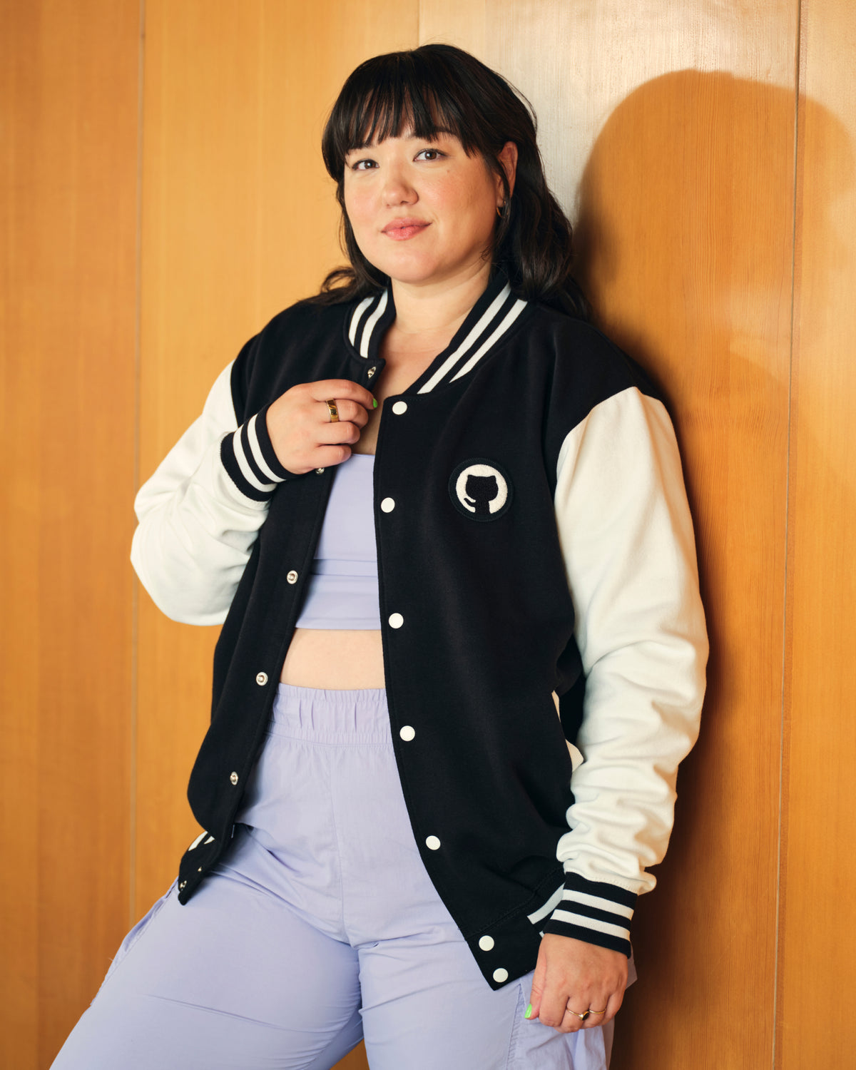 woman with black hair, posing wearing Mona Varsity Jacket over violet jumpsuit in office.