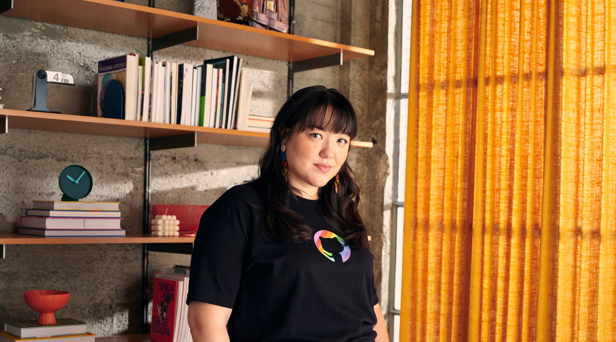 Model wearing a GitHub pride tee in an industrial room with a floating shelf and large curtained window background