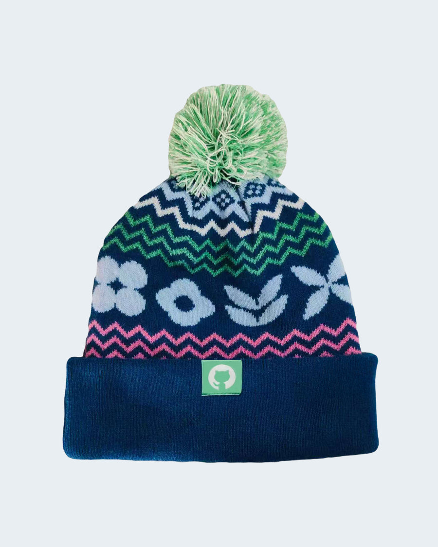 GitHub knit beanie with navy base, green zigzag, pink chevron, light blue floral design, and green pom-pom.