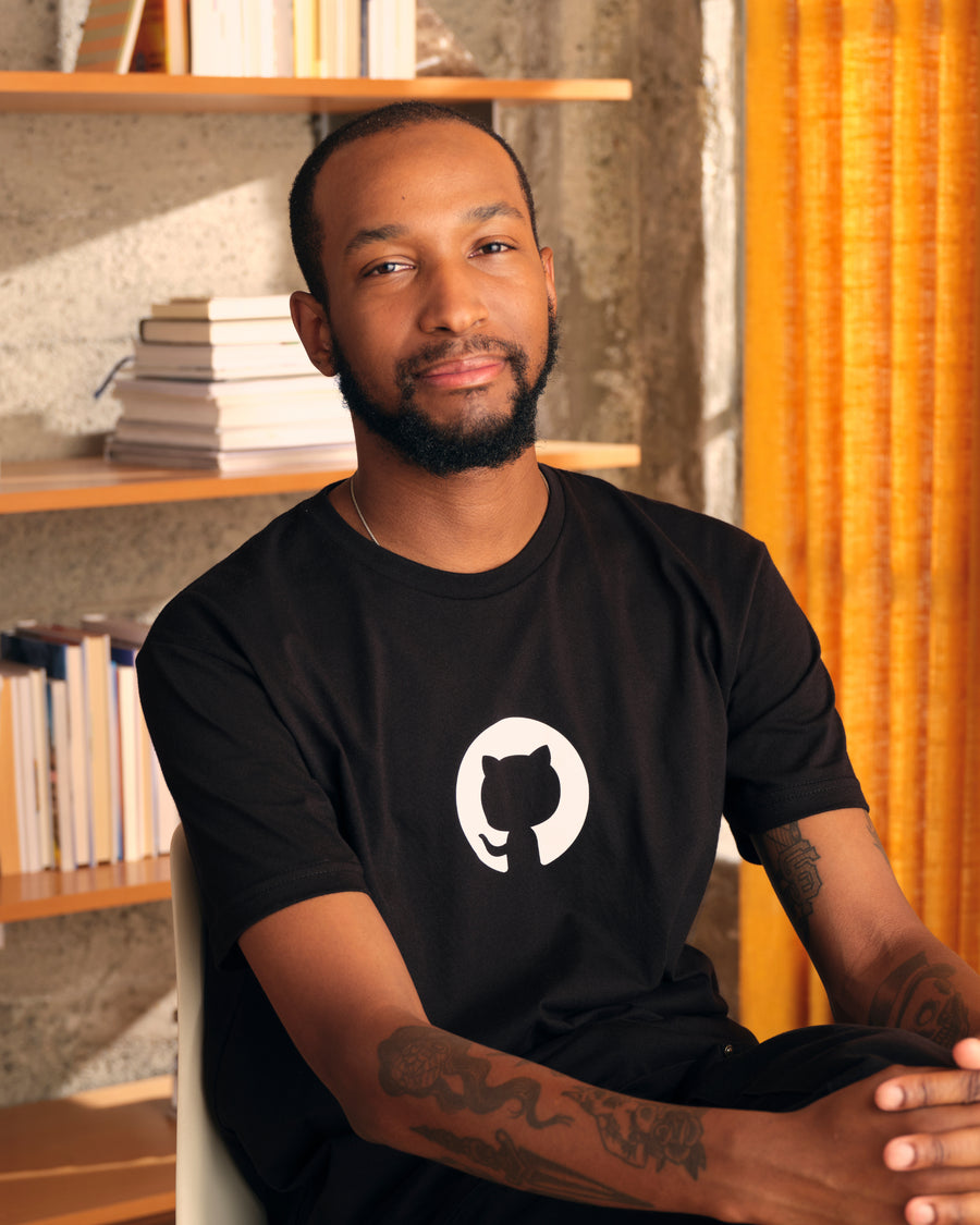 Photo of man, sitting while wearing  Invertocat 4.0 shirt in black in home office #color:black